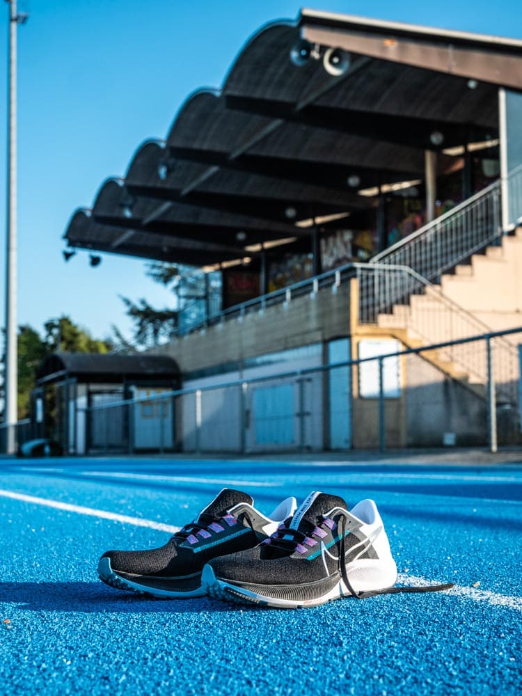 Paire de chaussure Nike Zoom Pegasus 38 posées sur le sol, sur une piste d'athlétisme en tartan bleu. Il y a une tribune en arrière plan.